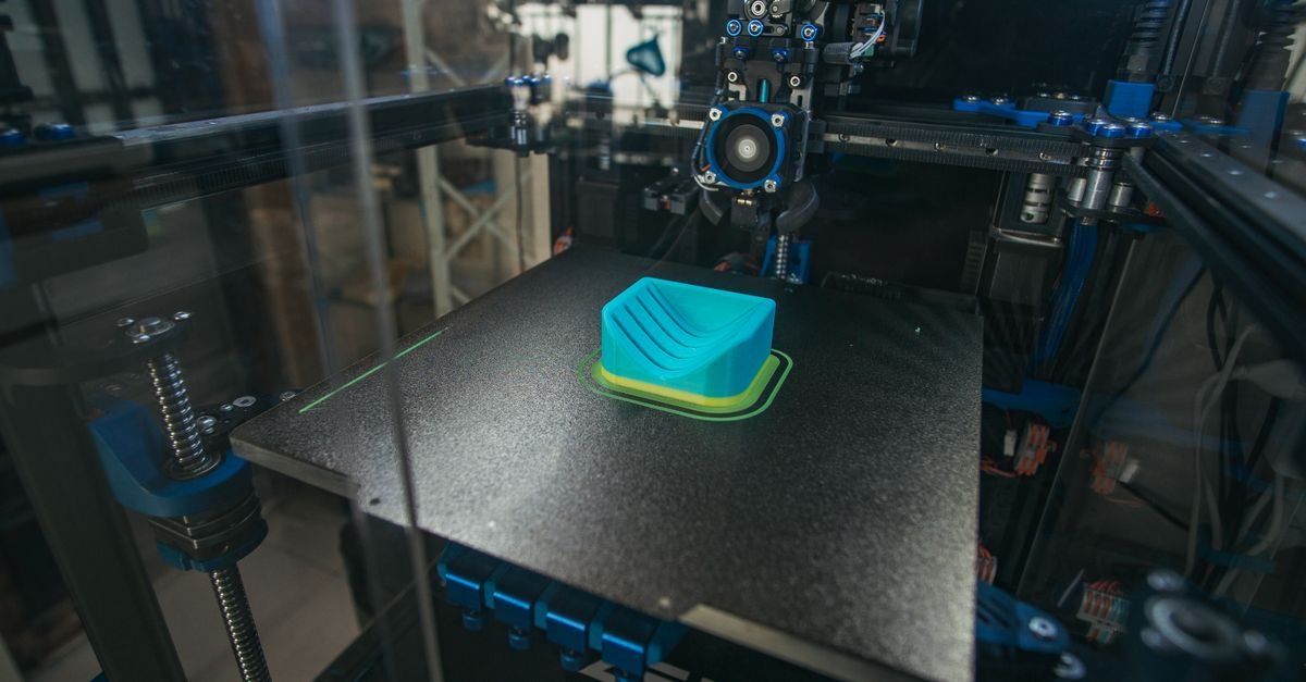 Impressora 3D produzindo uma peça plástica azul sobre a mesa de impressão