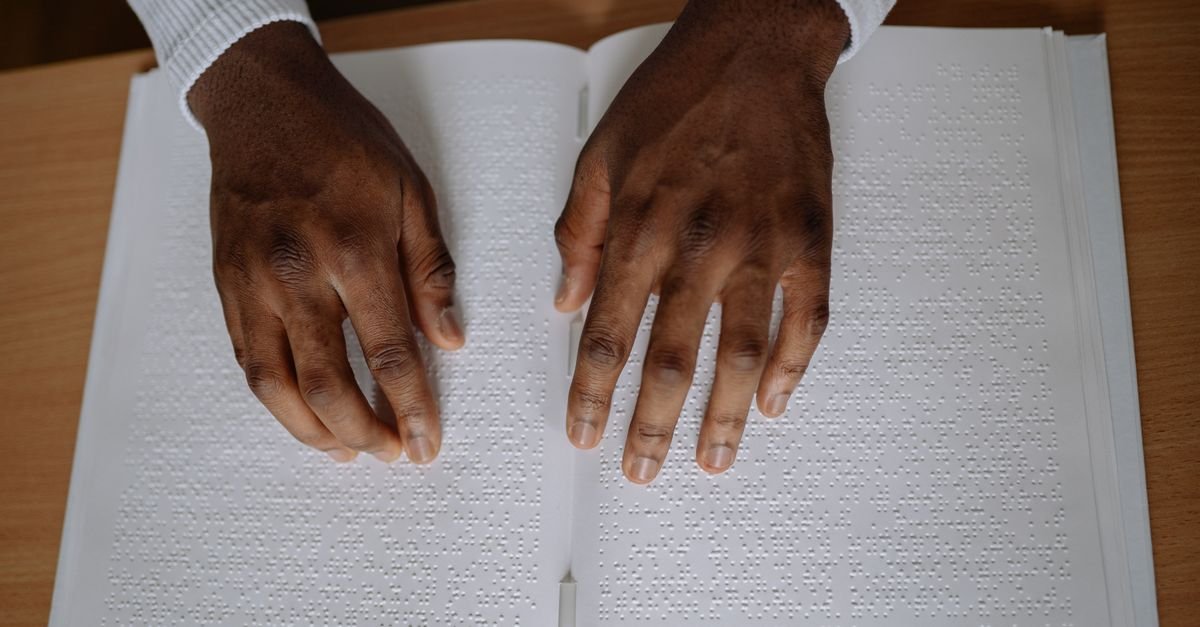 Mãos percorrendo as páginas de um livro em braile sobre mesa de madeira