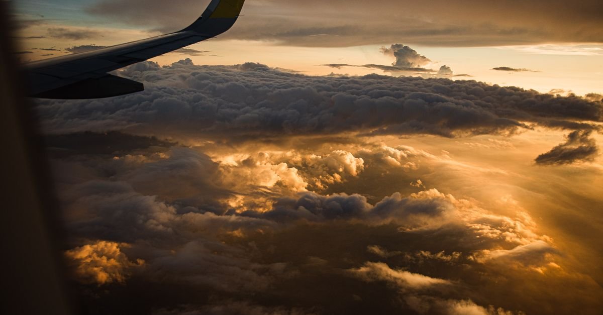 Asa de avião vista da janela durante o voo, acima de um mar de nuvens iluminado pelo pôr do sol