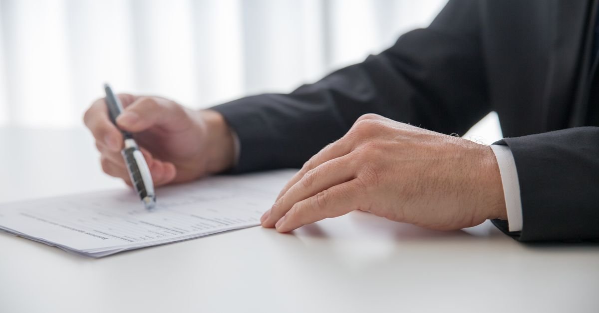 Mãos de homem de terno assinando um documento sobre a mesa