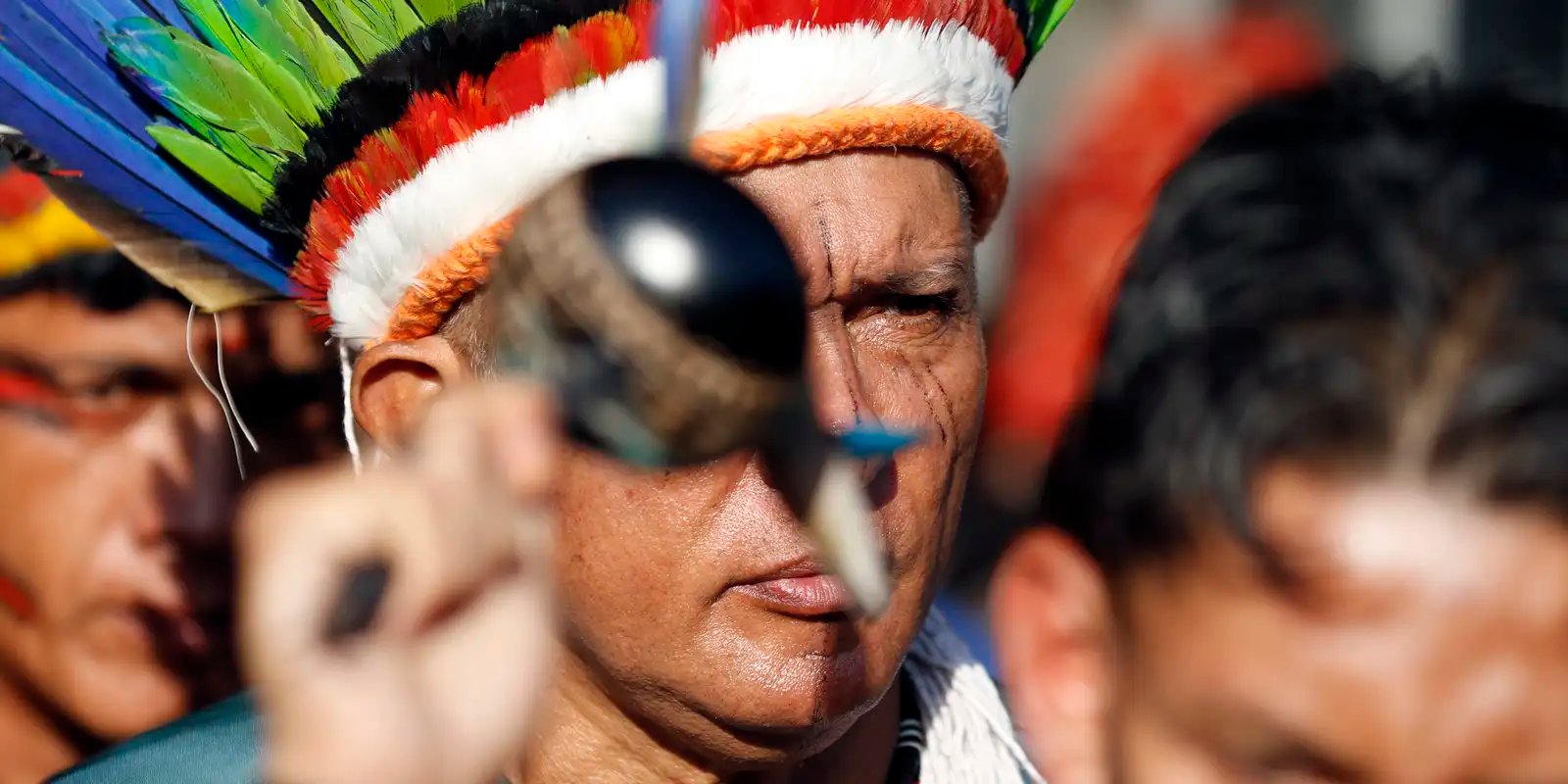 Homem indígena com cocar de penas coloridas em ato público, com outras pessoas ao fundo