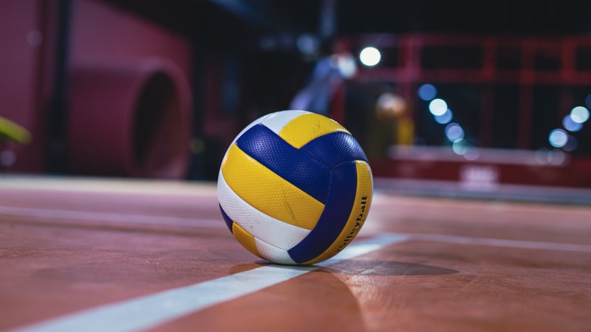Bola de vôlei azul, amarela e branca parada na linha de uma quadra coberta