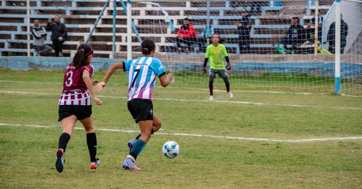 Jogadoras de futebol feminino disputam a bola em campo durante partida, com goleira ao fundo