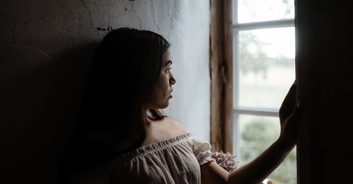 Mulher observa o lado de fora pela janela de uma casa, em ambiente de pouca luz