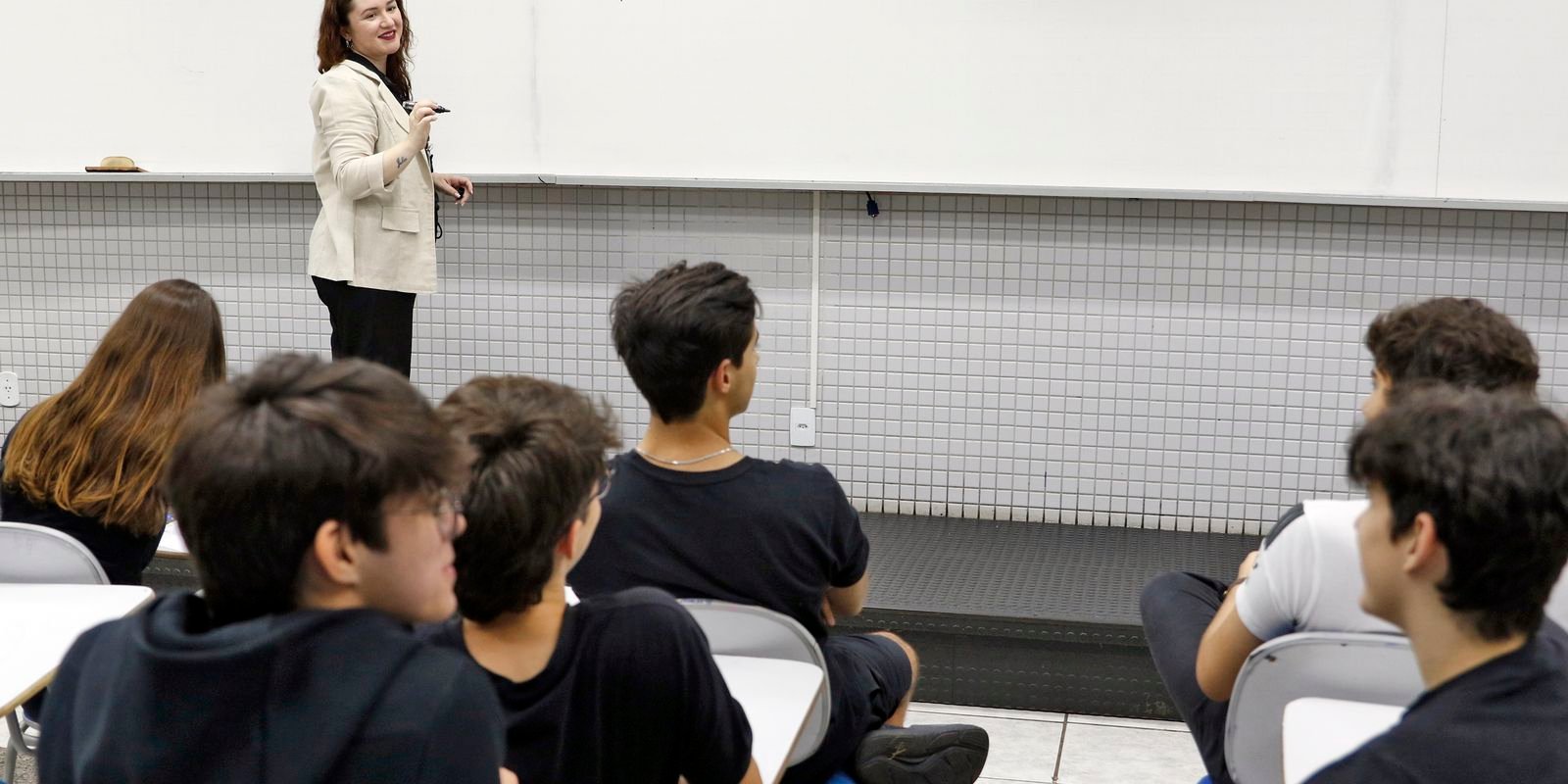 Professora em pé diante de quadro branco durante aula para estudantes sentados em carteiras