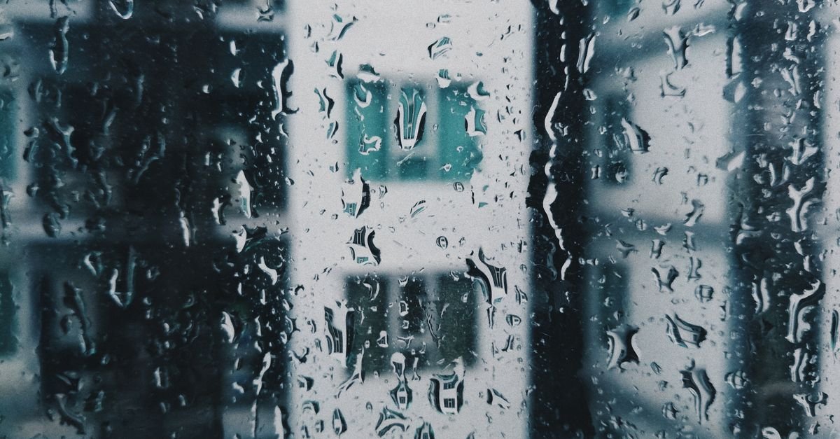 Gotas de chuva escorrem em vidro de janela com prédio desfocado ao fundo