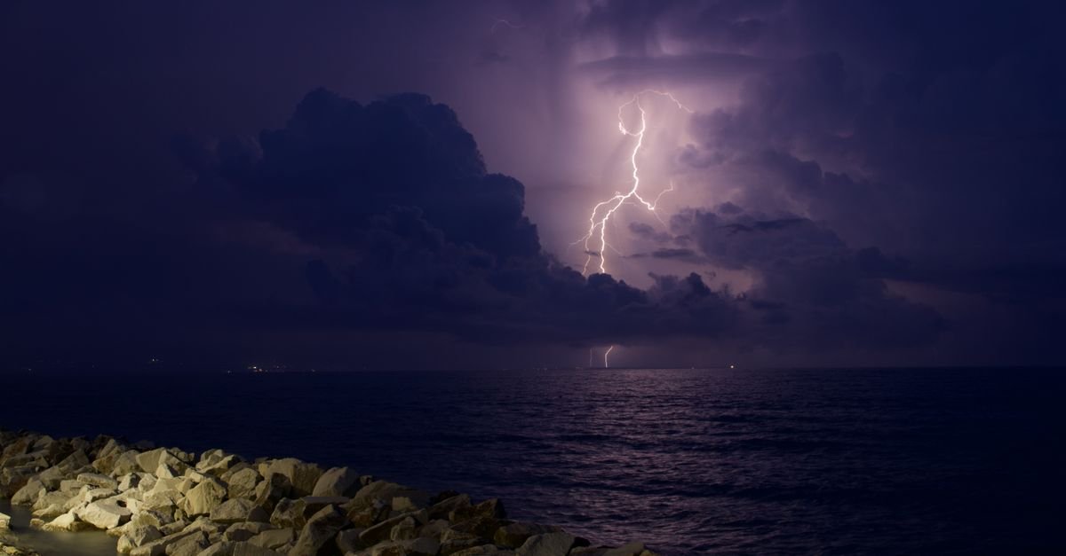 Raio cortando o ceu carregado sobre o mar durante tempestade noturna, com costao de pedras em primeiro plano