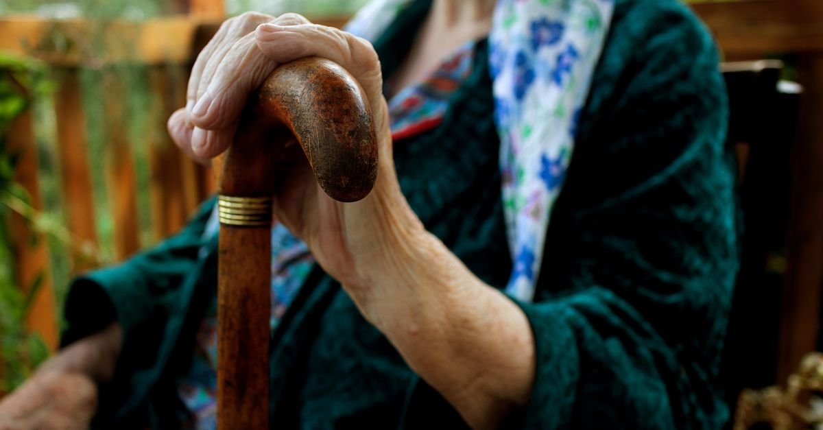 Mãos de uma pessoa idosa apoiadas sobre o castão de uma bengala de madeira