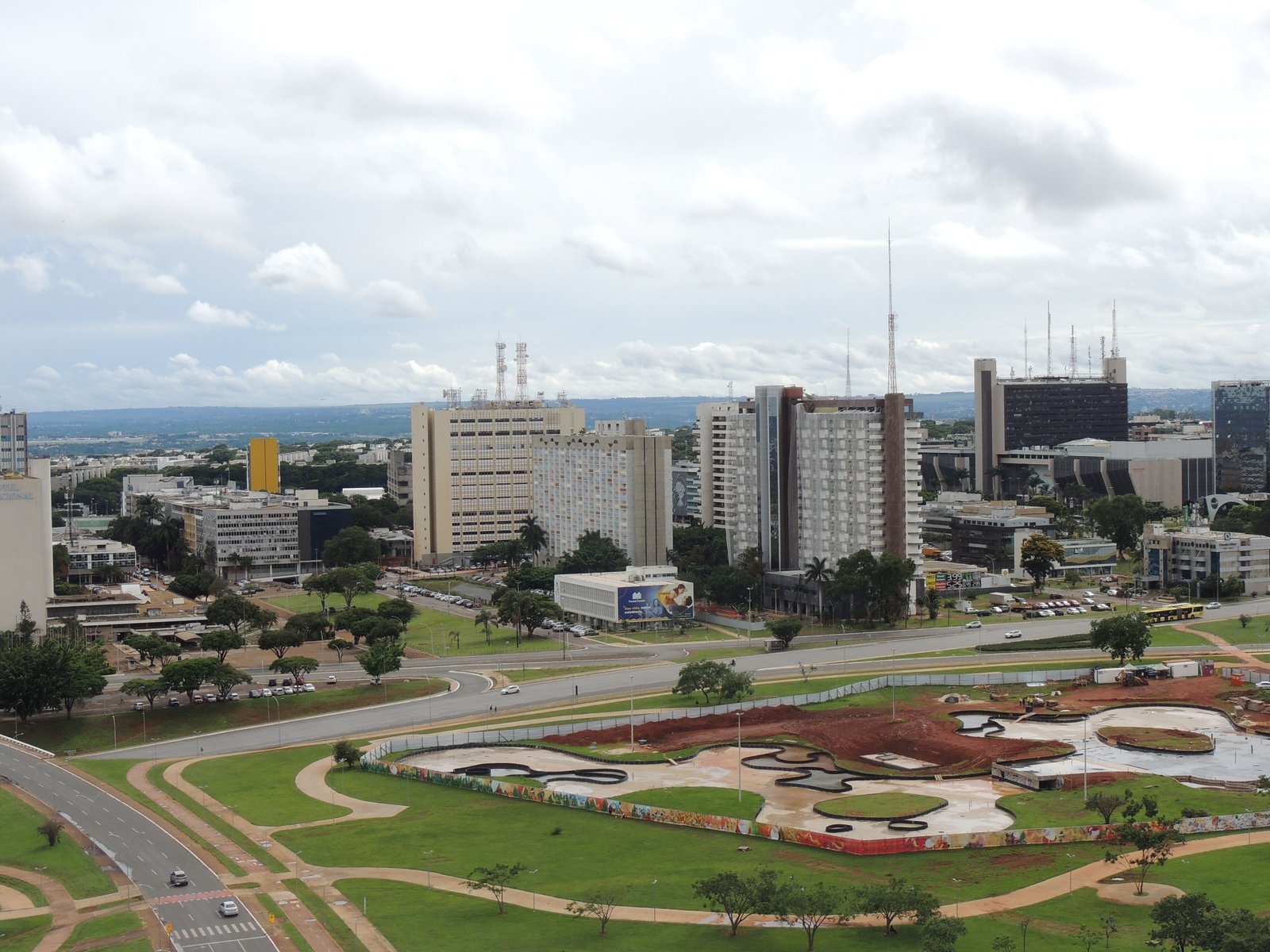 Vista elevada do Eixo Monumental, em Brasília, com prédios do setor comercial ao fundo e obras de urbanização em primeiro plano