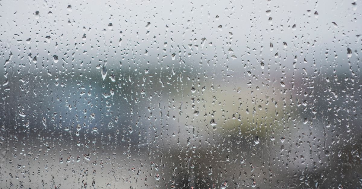 Gotas de chuva escorrem pelo vidro de uma janela, com a paisagem urbana desfocada ao fundo