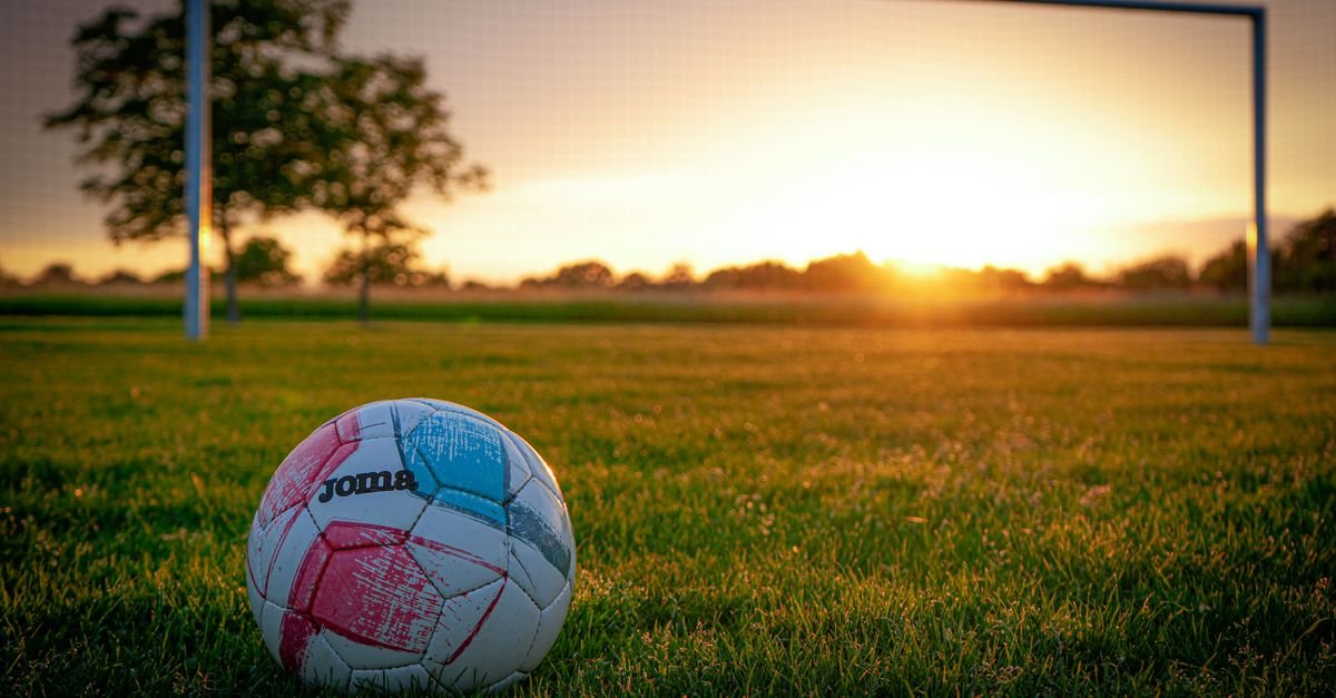A soccer ball on a grassy field with a goal post, beautifully lit by the vibrant sunset.