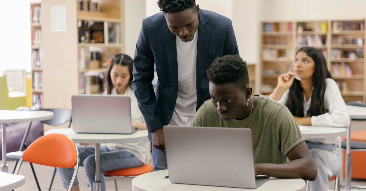 Students learning in a classroom setting with a teacher assisting and laptops on desks, creating an interactive education environment.