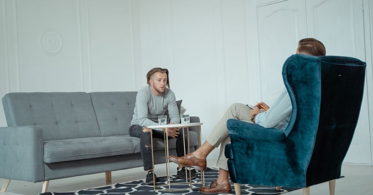 Two men engaged in a therapy session inside a modern office setting, discussing mental health.