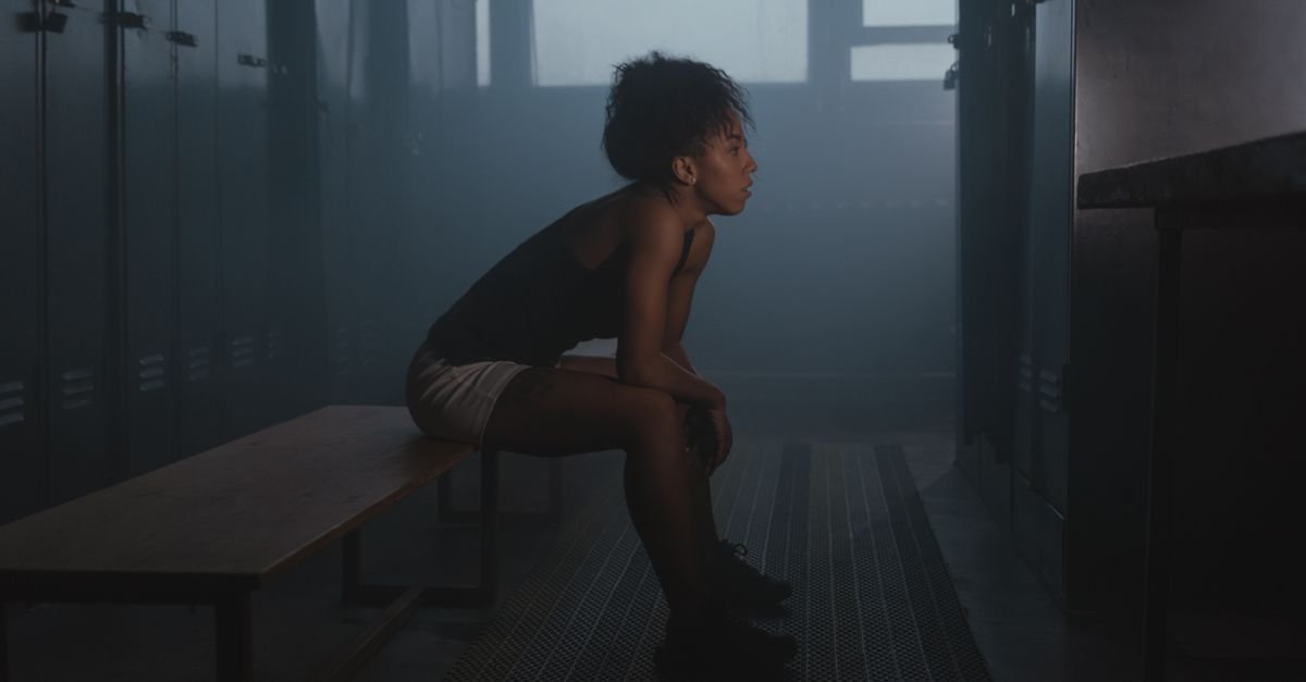 Woman sitting on a bench in a dimly lit locker room, exuding a pensive mood.
