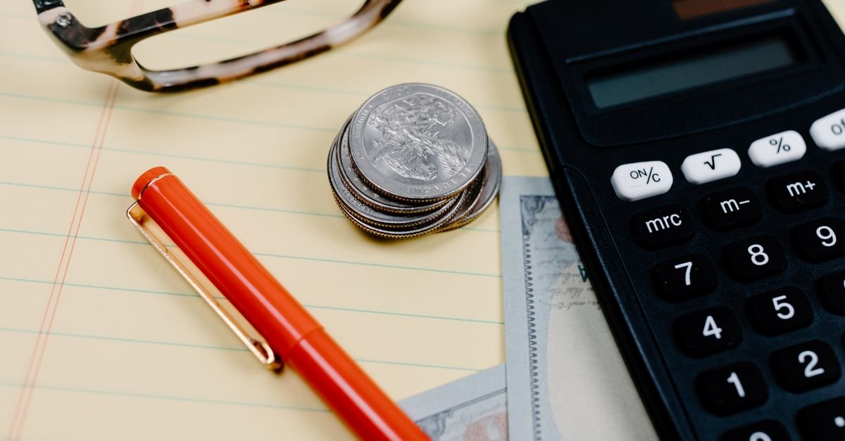 Close-up of a workspace with a calculator, coins, and glasses on a notepad.