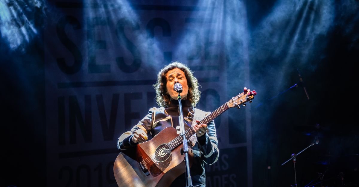 Musician performing live with guitar at Teresópolis festival, stage lit with colorful lights.