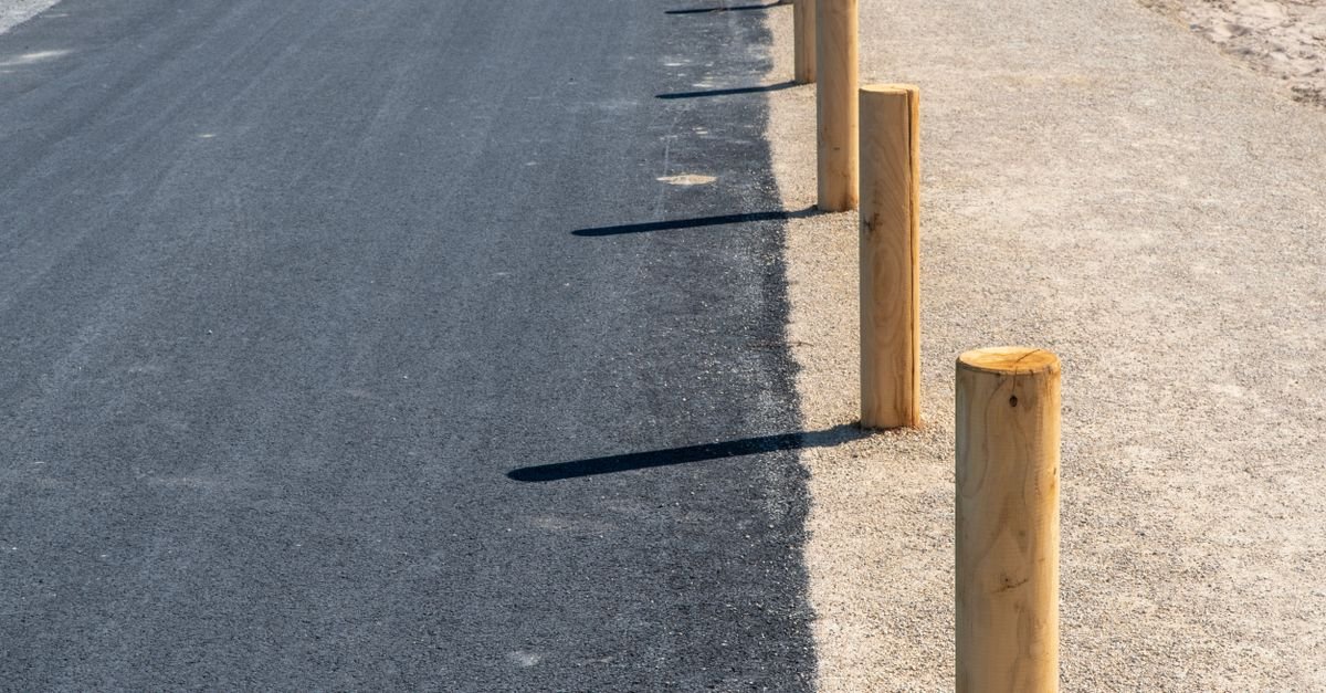A freshly paved asphalt road bordered by sandy path and wooden posts in bright sunlight.