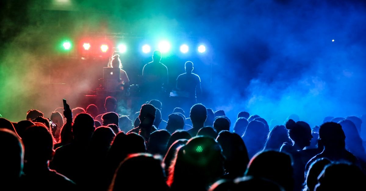 Silhouettes of a lively crowd enjoying a DJ's performance with colorful stage lights at night.