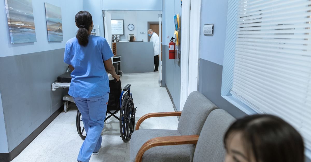 A nurse in scrubs pushes a wheelchair through a well-lit hospital corridor, attending to patient care.