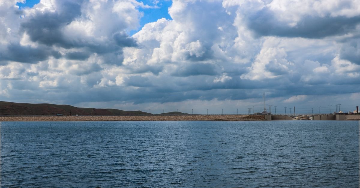 Expansive reservoir with dramatic clouds and distant structures under a bright blue sky.