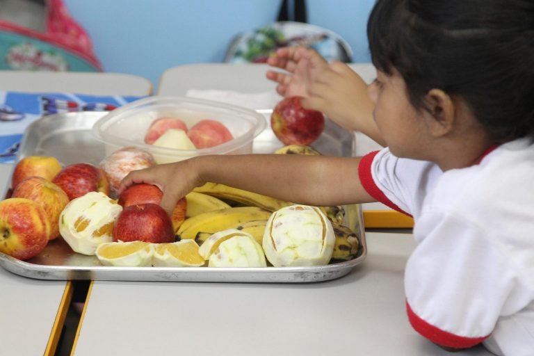 Criança pega fruta em bandeja com maçãs, bananas e laranjas em escola