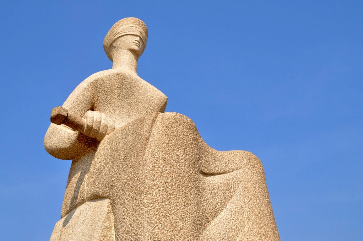 Estátua da Justiça, escultura de mulher de olhos vendados com espada sobre os joelhos, contra o céu azul