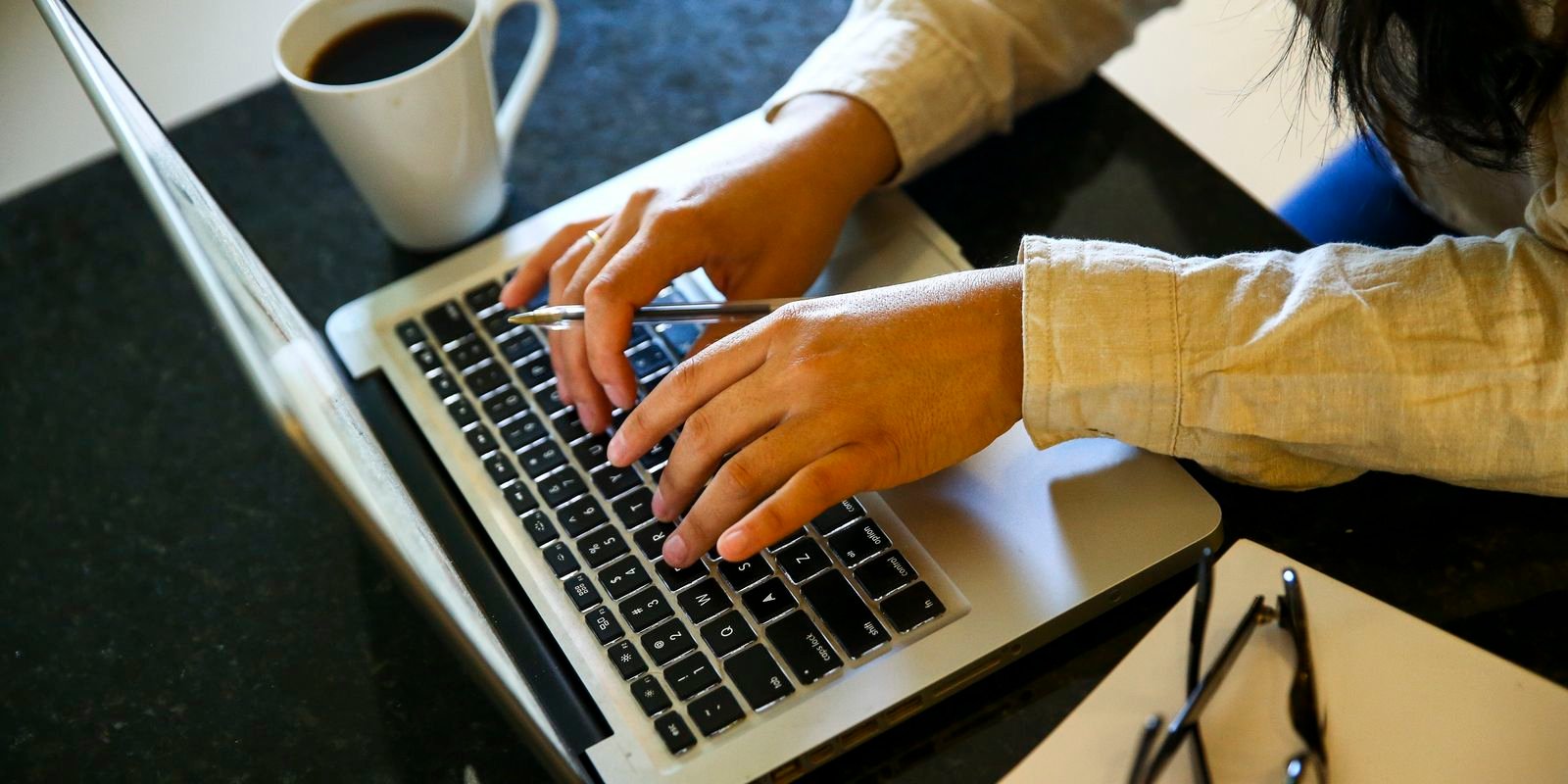 Pessoa digita em notebook sobre mesa de trabalho, com xícara de café ao lado