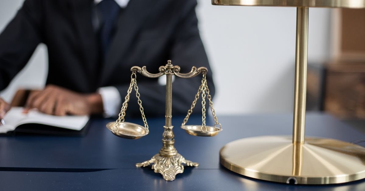 Balança da justiça dourada sobre mesa de escritório, com pessoa desfocada ao fundo