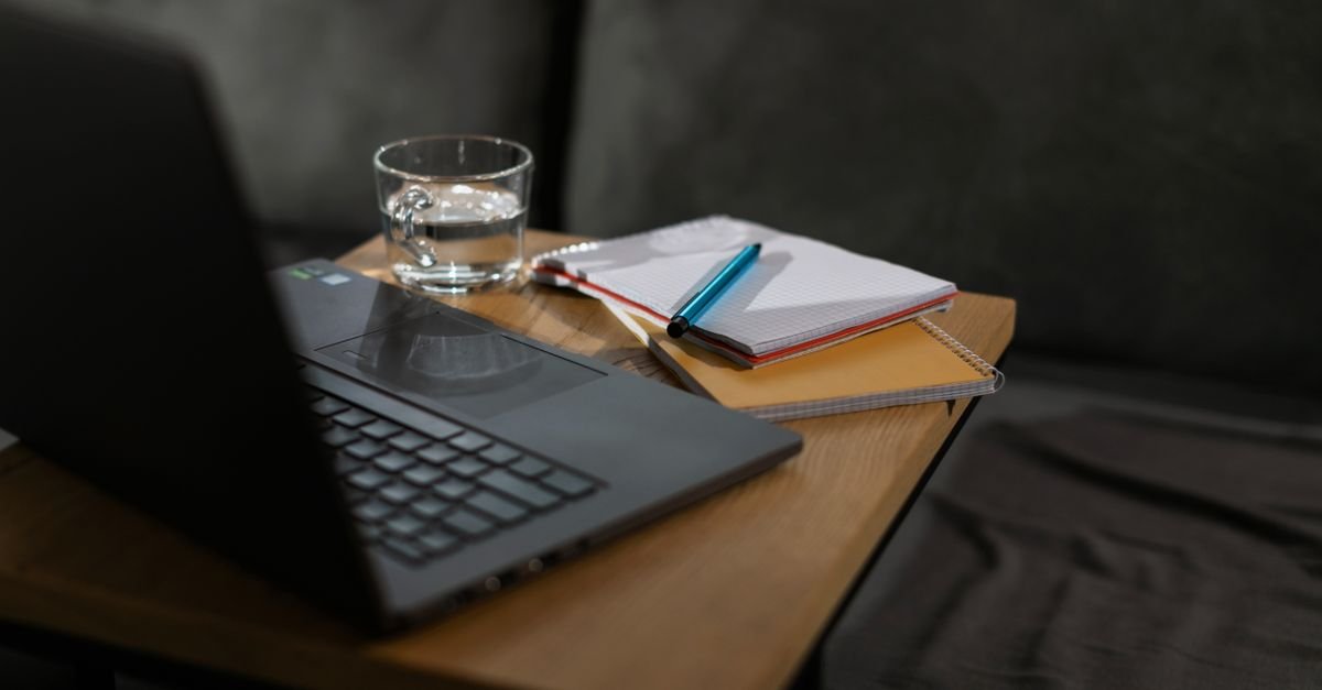 Notebook aberto, cadernos e uma caneta sobre mesa de madeira em ambiente doméstico, representando o estudo em casa