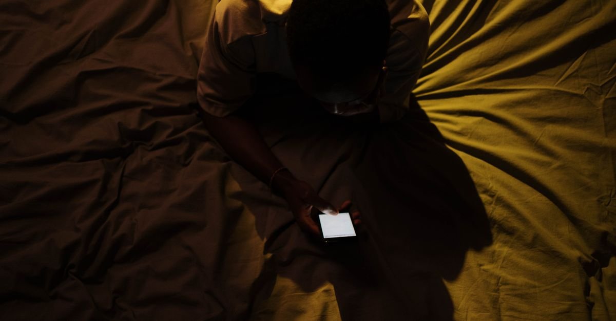 Pessoa vista de cima usando smartphone em um quarto escuro, com a tela iluminando as mãos