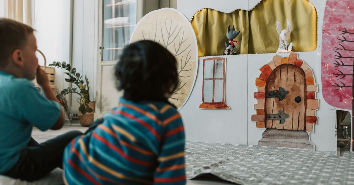 Duas crianças pequenas sentadas no chão assistindo a um teatro de fantoches montado em casa