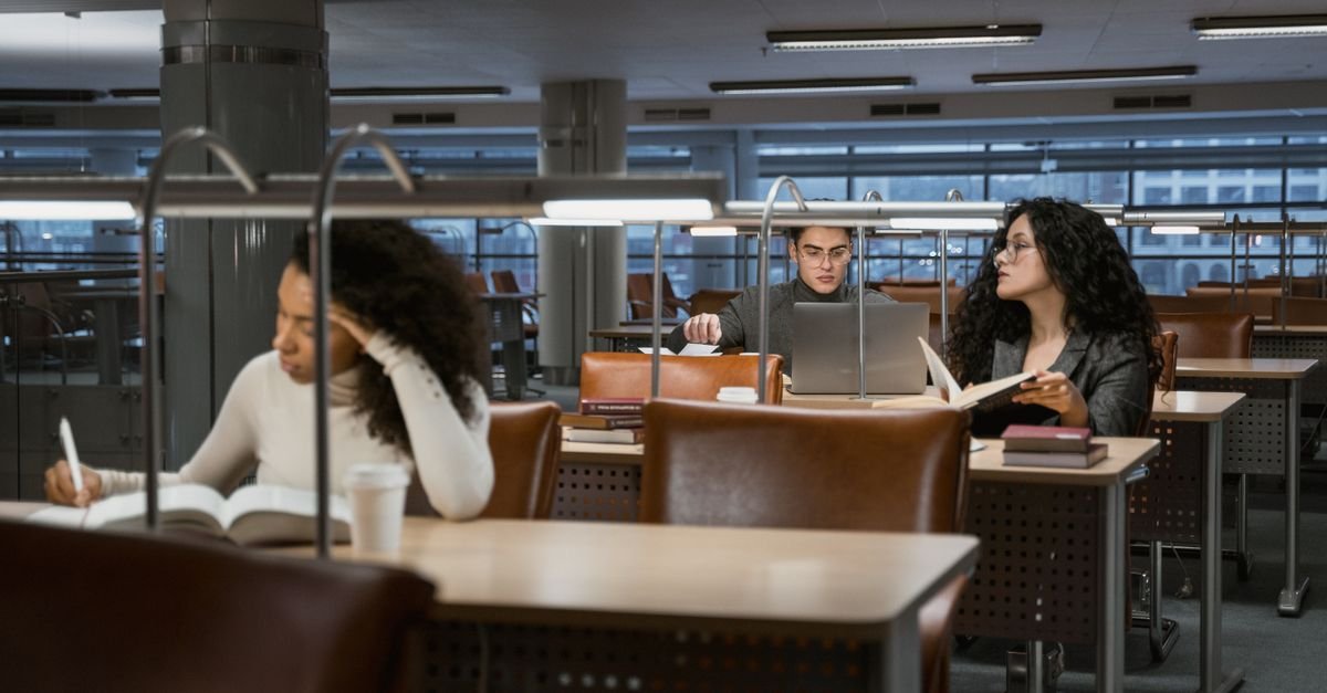 Estudantes estudando com livros e notebook em mesas de biblioteca universitária