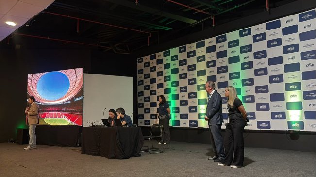 Abertura da inspeção da Fifa em sala do Mané Garrincha, com imagem aérea do estádio projetada em telão e painel com a marca da Arena BRB ao fundo