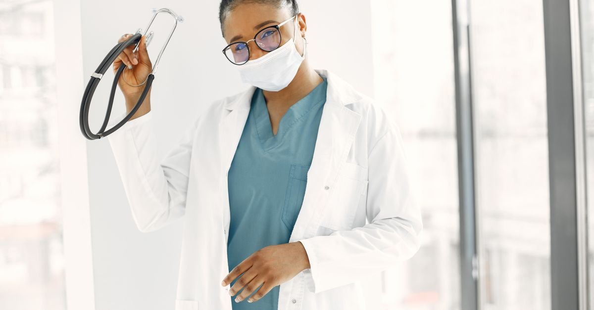 Médica de jaleco branco e máscara segura um estetoscópio em corredor iluminado de unidade de saúde