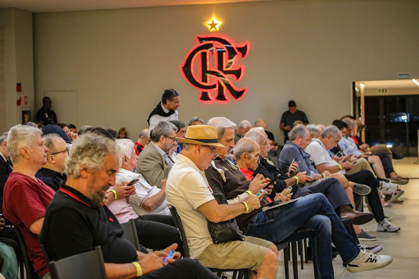 Conselheiros do Flamengo sentados durante sessão do Conselho Deliberativo, com o escudo do clube iluminado ao fundo