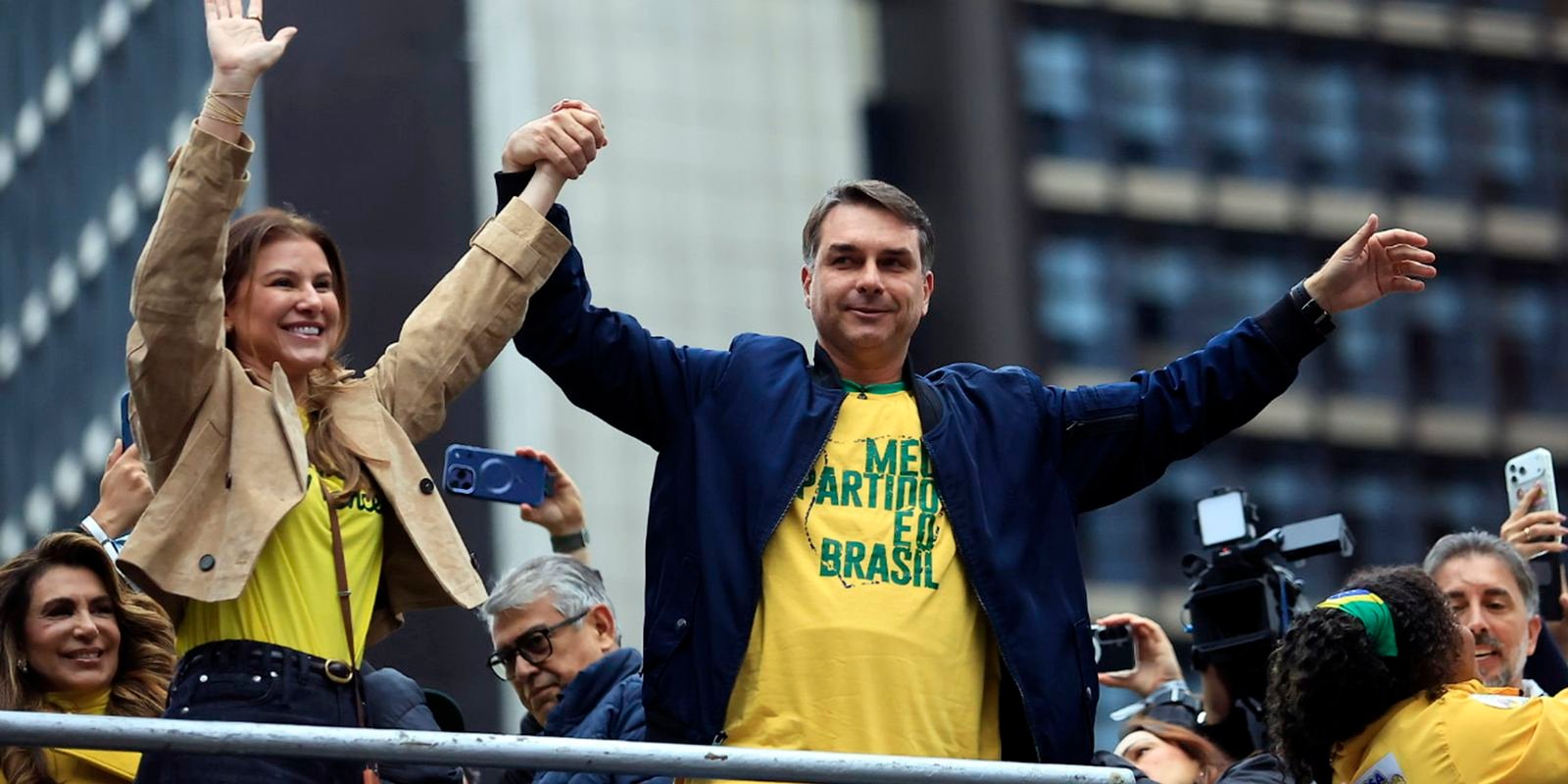 Flávio Bolsonaro de braços erguidos durante ato de campanha, ao lado da mulher, com apoiadores de camiseta amarela ao redor