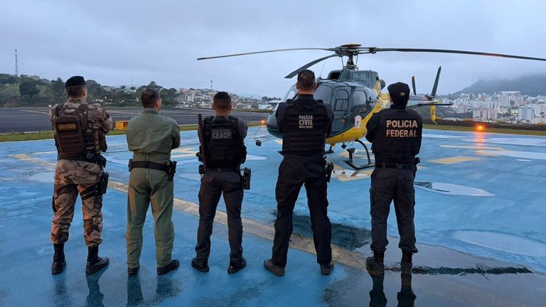 Cinco policiais de costas diante de um helicóptero da Polícia Federal em heliponto, com coletes identificados de Polícia Federal, Polícia Civil e Ceaop