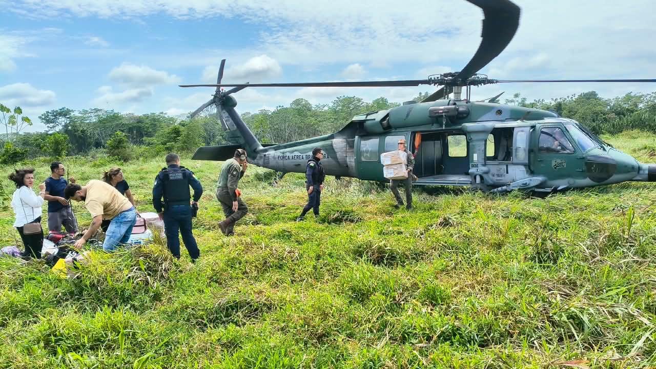 Helicóptero da Força Aérea Brasileira pousado em campo aberto enquanto militares descarregam caixas e eleitores esperam ao lado