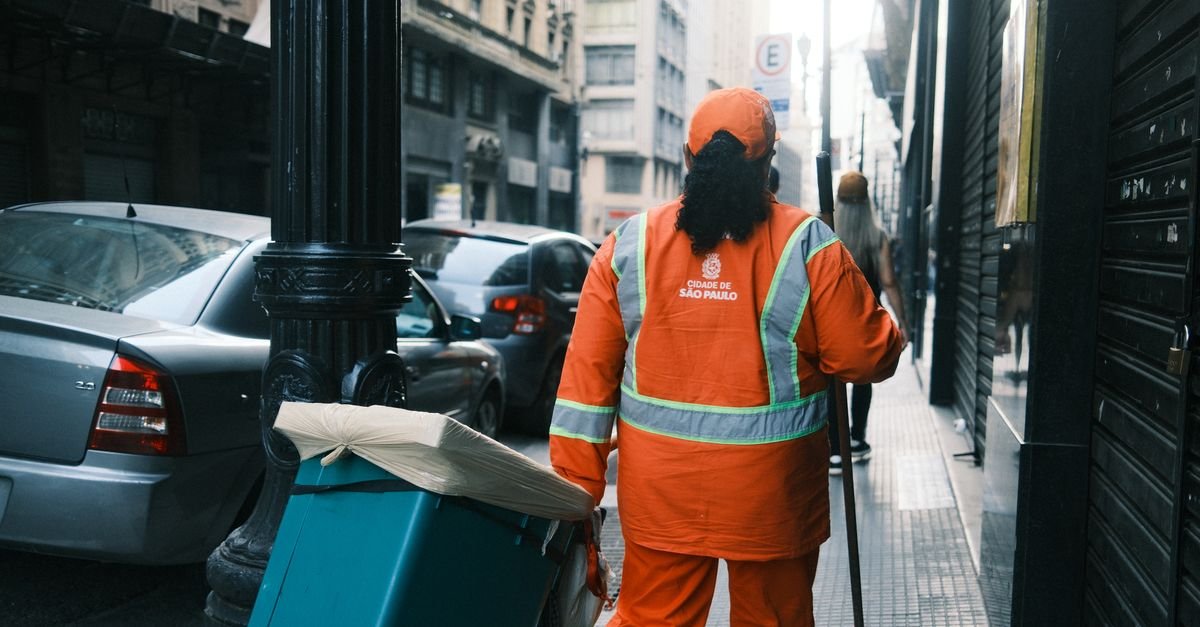 Gari de uniforme laranja da cidade de São Paulo empurra contêiner de lixo em calçada do centro