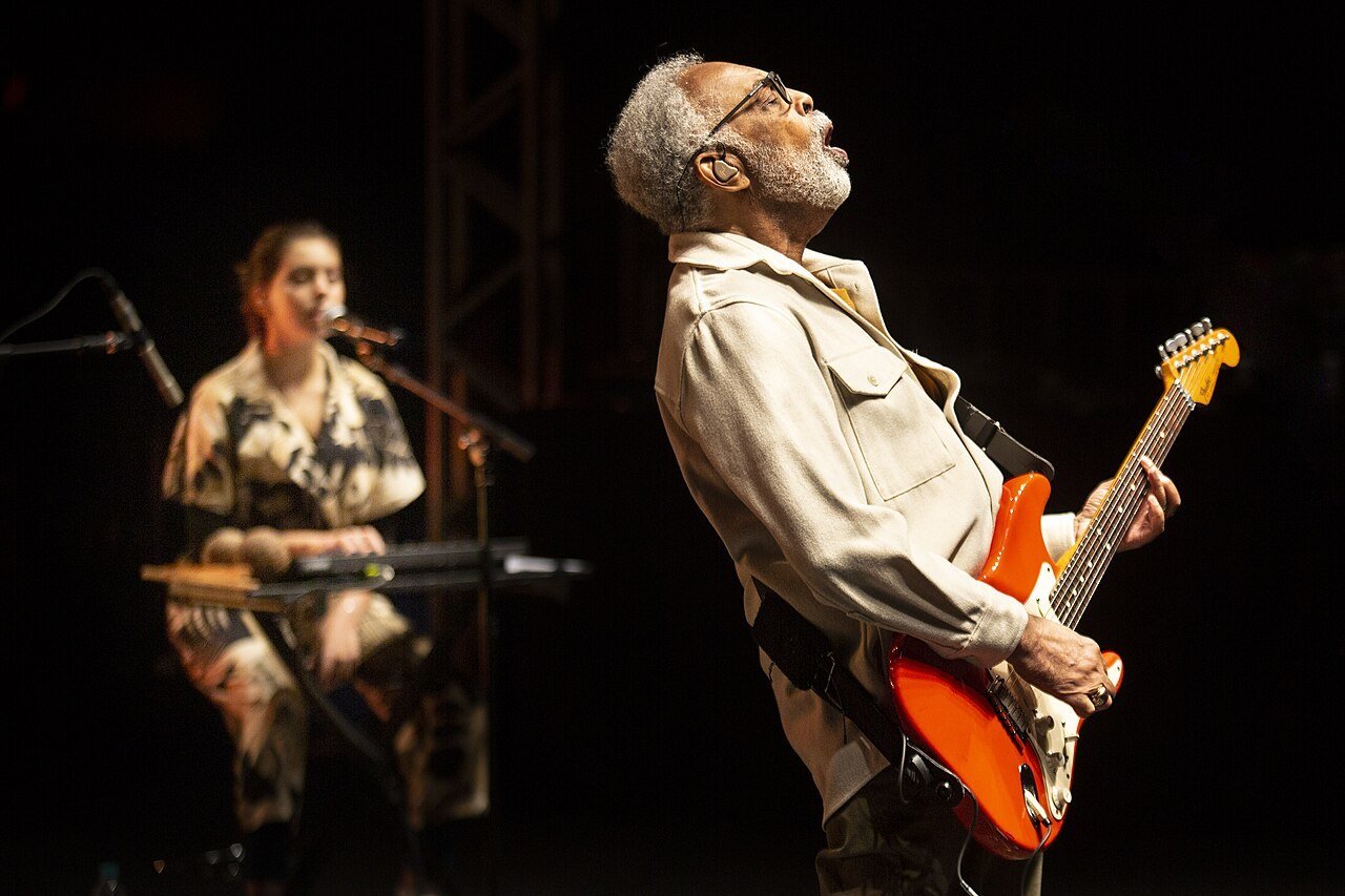 Gilberto Gil canta no palco com guitarra laranja, de cabeça erguida, sob luz quente