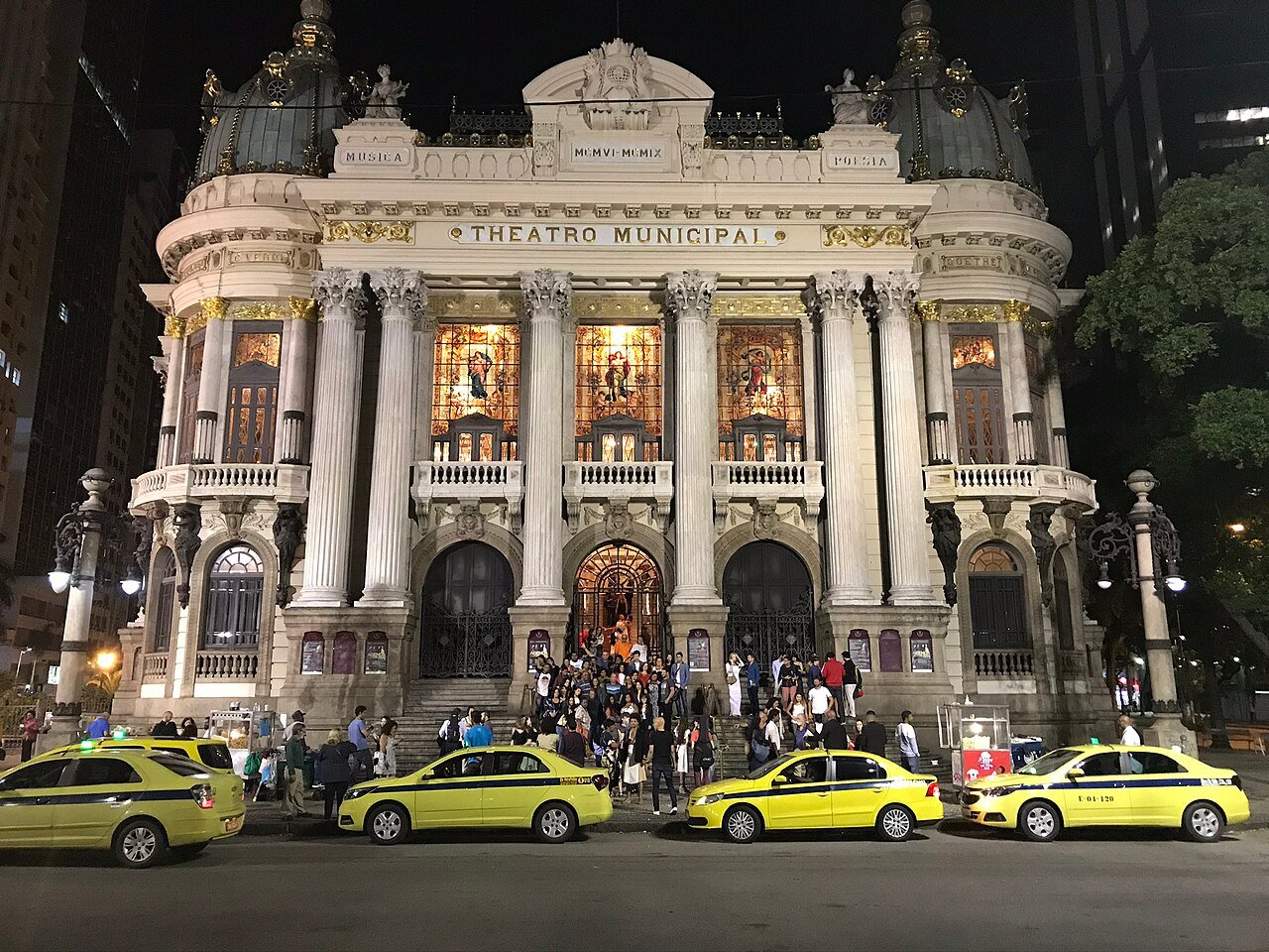 Fachada iluminada do Theatro Municipal do Rio de Janeiro à noite, com público na escadaria e táxis estacionados em frente
