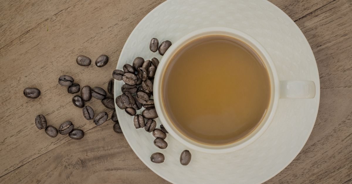 Xícara branca com café vista de cima, com grãos espalhados sobre a mesa
