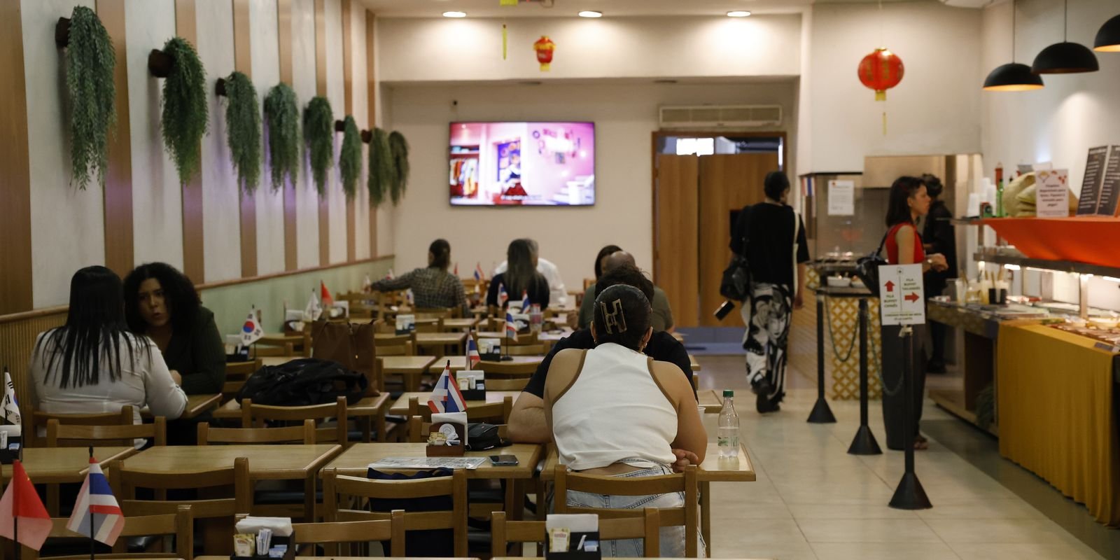 Clientes sentados em mesas de restaurante self-service com balcão de bufê ao fundo