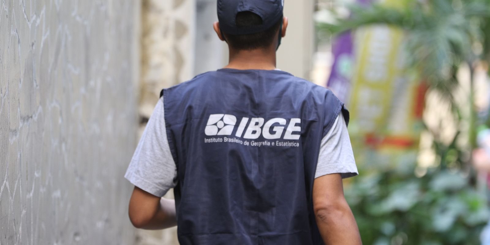 Recenseador do IBGE de costas, com colete de identificação do instituto, durante trabalho de campo