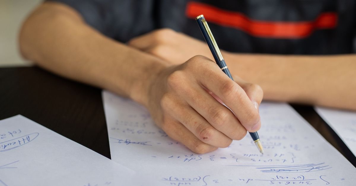 Mão de estudante escrevendo equações em folha de papel sobre uma mesa
