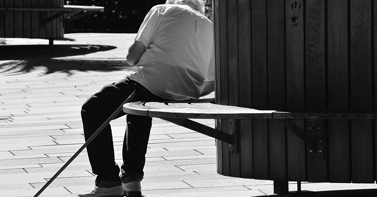 Homem idoso sentado em banco de praça, de costas, apoiado em uma bengala, em foto preto e branco