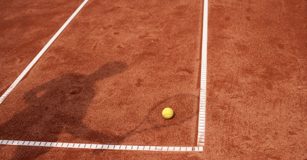 Bola de tênis parada sobre a linha de uma quadra de saibro, com a sombra de um jogador ao lado