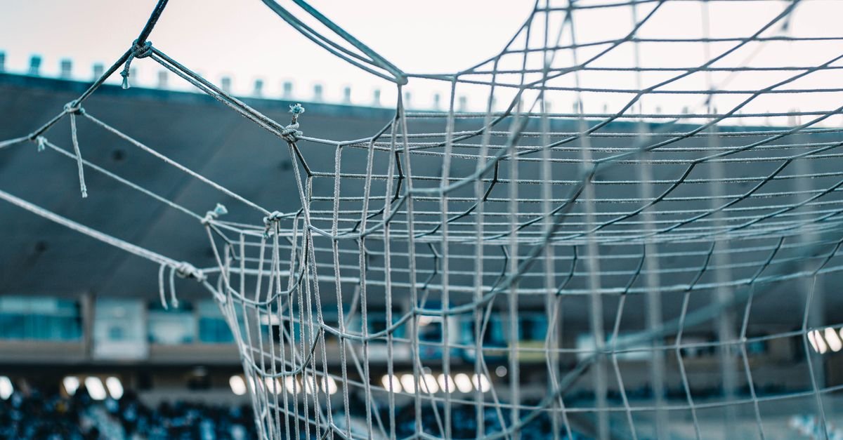Rede de gol de futebol em primeiro plano, com as arquibancadas vazias de um estádio ao fundo