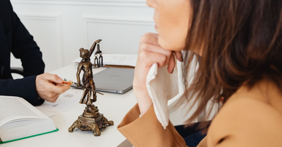 Estátua de bronze da Justiça sobre mesa branca, com pessoas conversando ao fundo