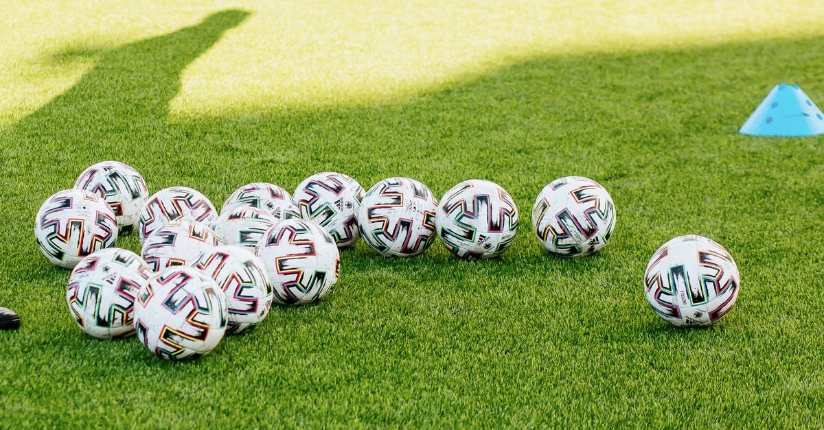 Bolas de futebol espalhadas sobre gramado sintético ao lado de um cone de treino